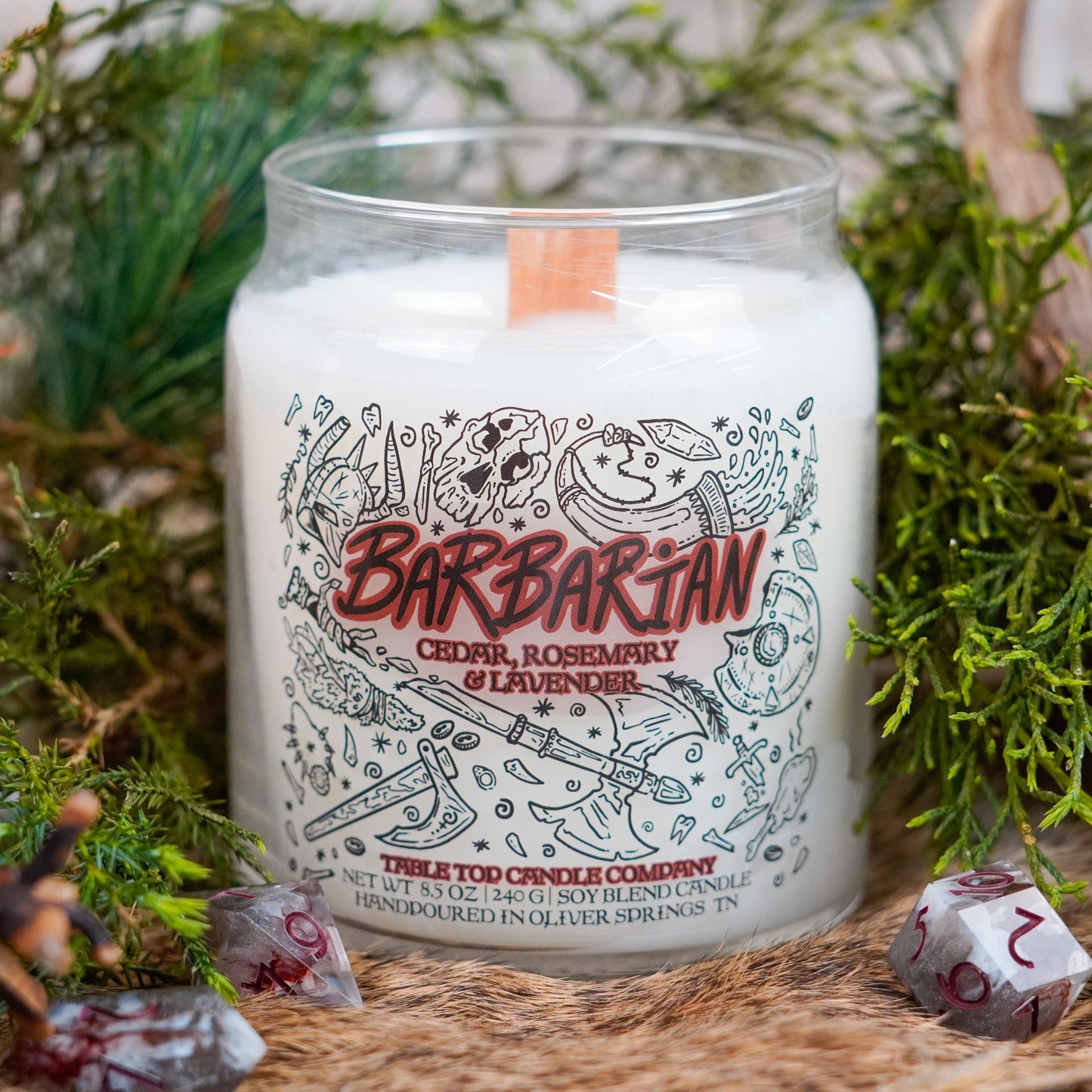 A medium-sized white soy candle labeled "Barbarian" with the scent cedar, rosemary, and lavender. The candle features fantasy-themed illustrations and a wooden wick, surrounded by polyhedral dice and forest greenery.
