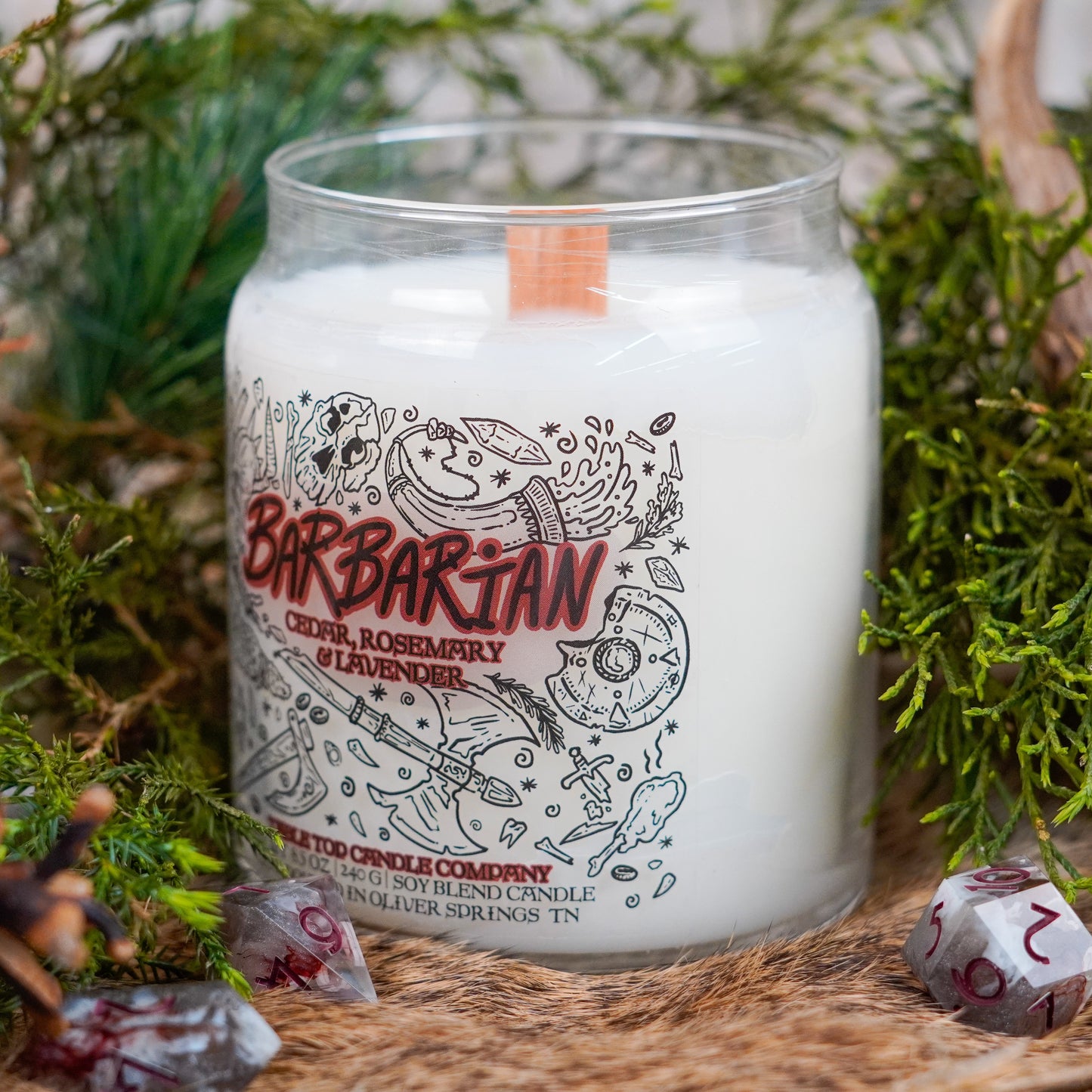 A side-angled view of the "Barbarian" candle by Table Top Candle Company, showing its detailed fantasy artwork label and wooden wick. The candle is nestled among faux fur, pine needles, and polyhedral dice.
