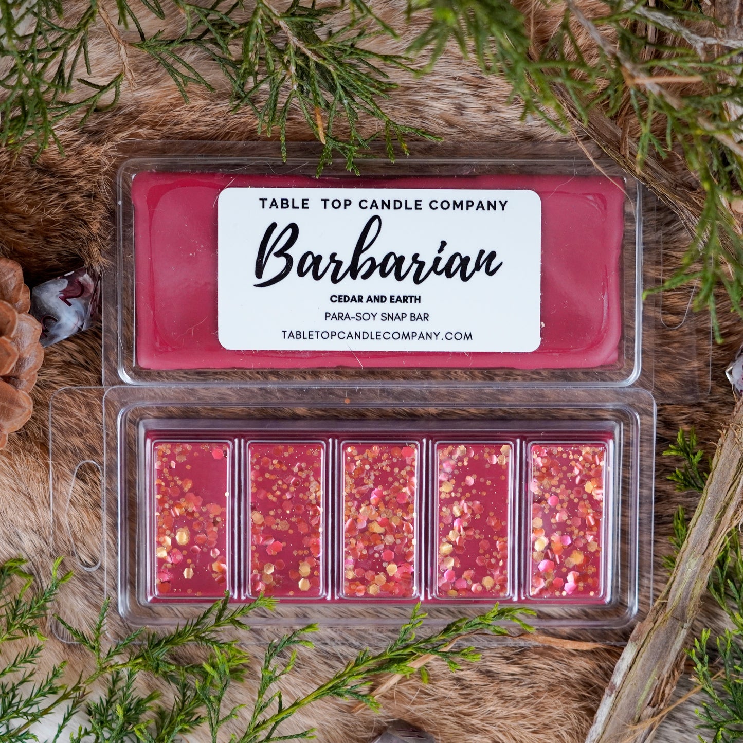 A top-down view of a red para-soy snap bar labeled "Barbarian" from Table Top Candle Company. The scent is Cedar and Earth. The wax bar features five glitter-filled segments and is displayed on a fur surface with pinecones, branches, and greenery for a rustic, fantasy-inspired aesthetic.