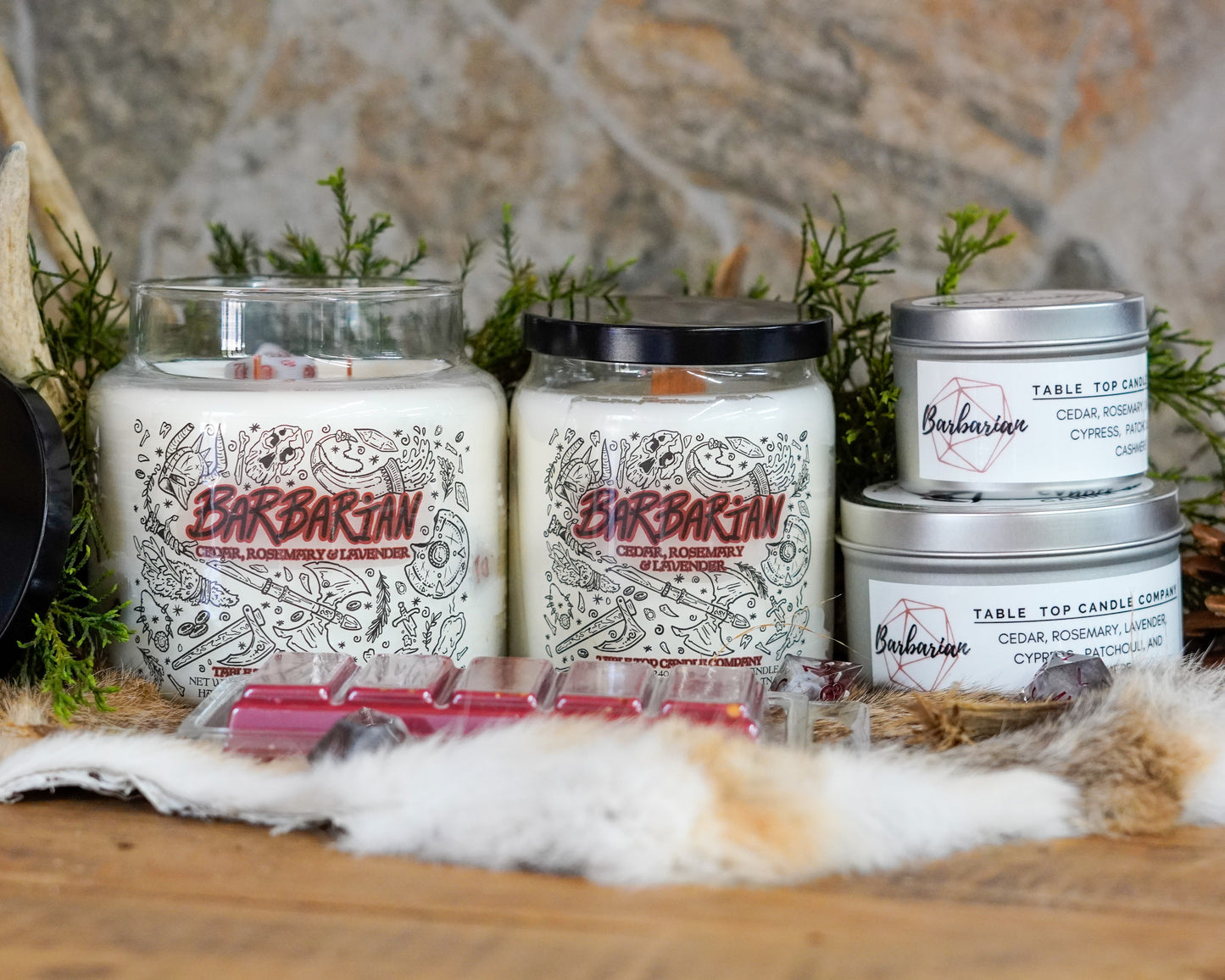 A product lineup of the "Barbarian" candle collection by Table Top Candle Company, including large and small jar candles, tins, and wax melts. All products feature the cedar, rosemary, and lavender scent, displayed on a wood and fur backdrop with natural greenery.