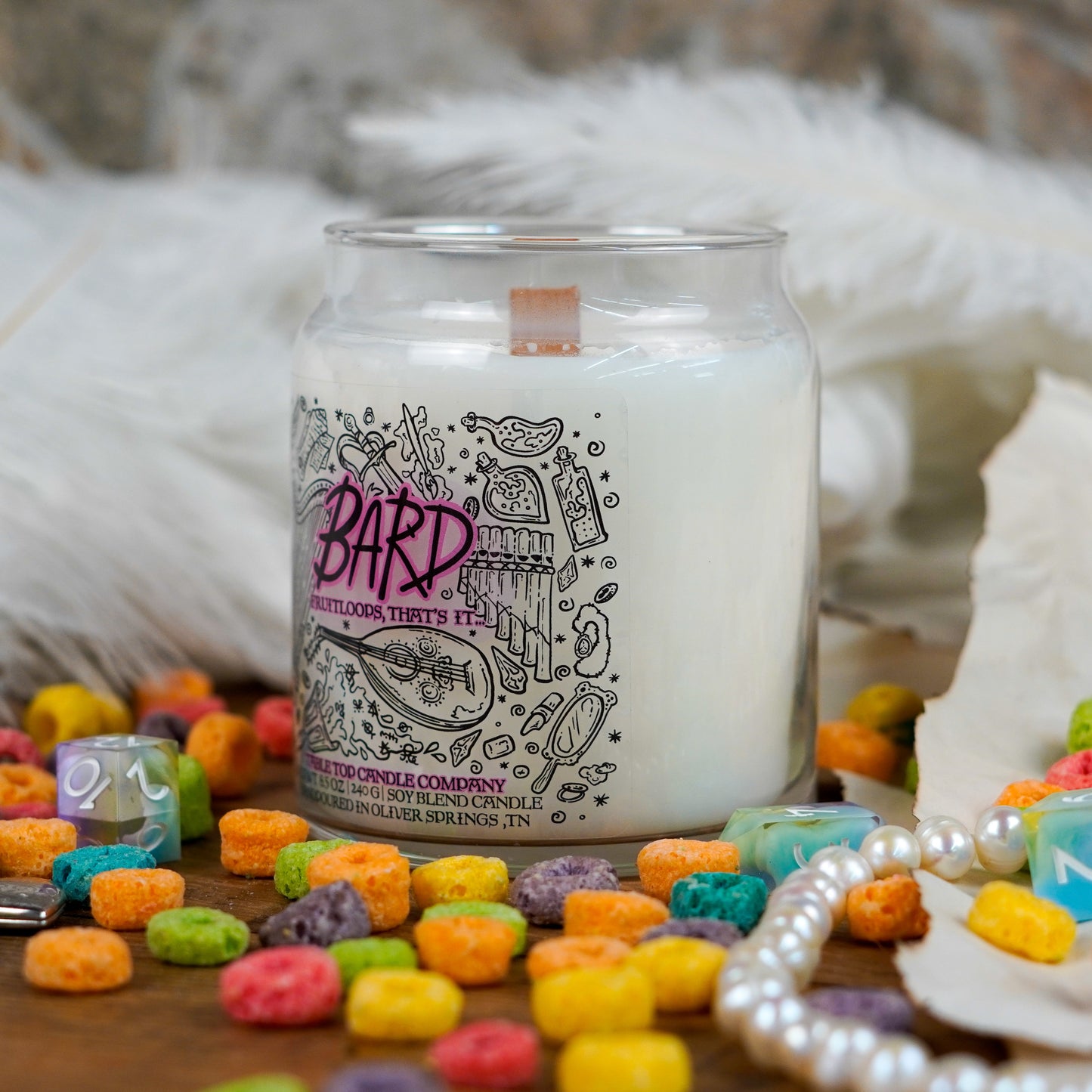 Angled view of the "Bard" candle highlighting its label and wooden wick. Set in a whimsical flat lay with fruit loop cereal, a pearl necklace, and feathers.