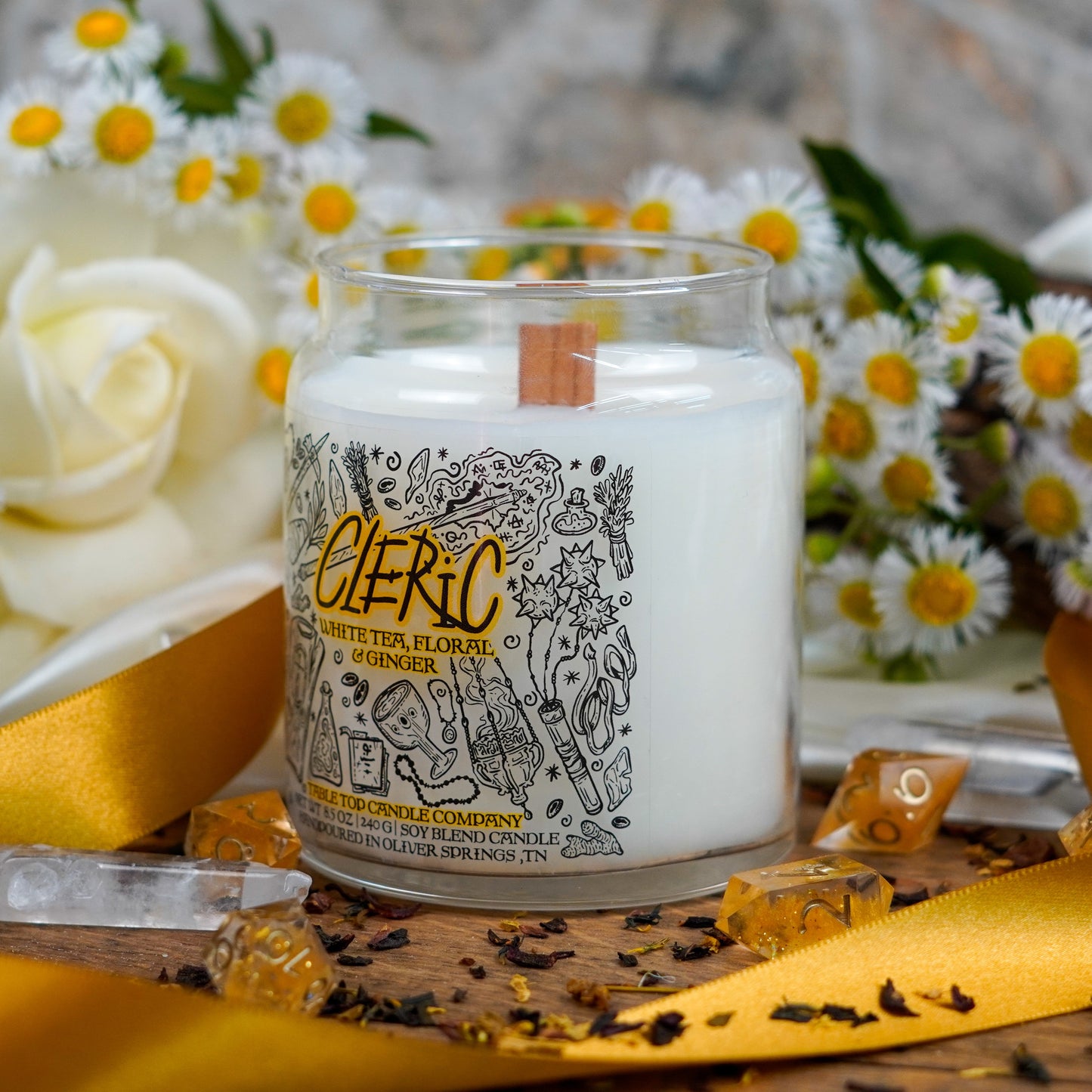 Angled close-up of the "Cleric" candle highlighting its illustrated label and wooden wick. The setting includes tea leaves, crystal points, and cheerful daisy flowers.