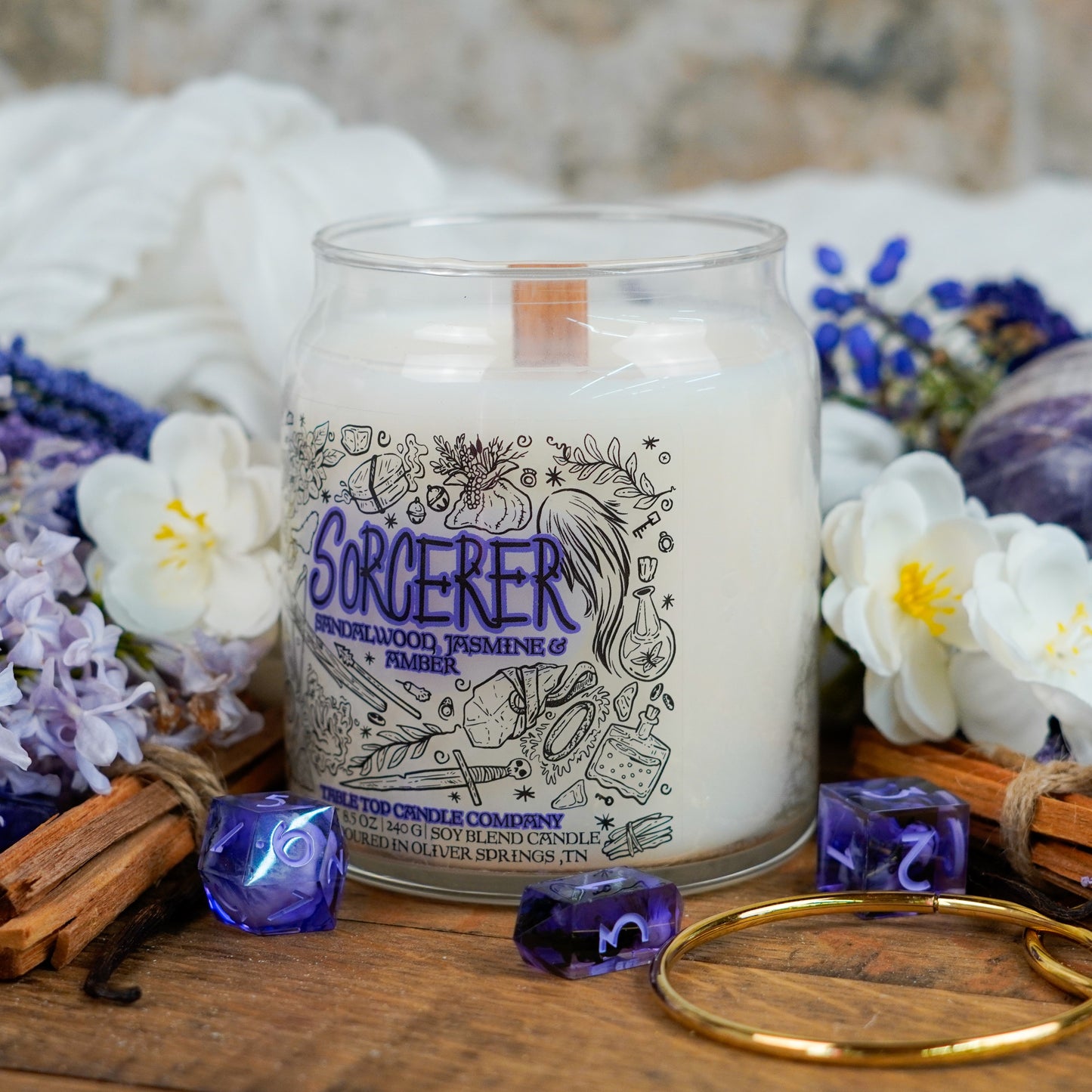 Angled side view of the "Sorcerer" candle with soft lighting, surrounded by floral arrangements and dice. The label highlights spellcasting tools and ingredients in a fantasy-inspired design.