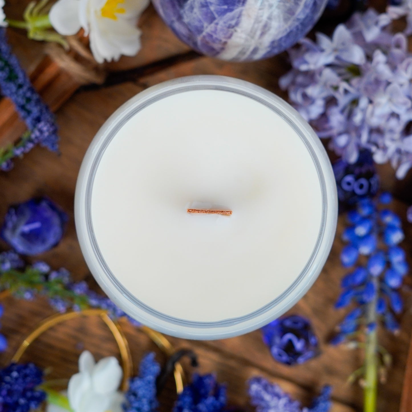 Overhead shot of the unlit "Sorcerer" candle, showing a single wooden wick in creamy white wax. The setup includes purple dice, florals, and a cozy wooden background.