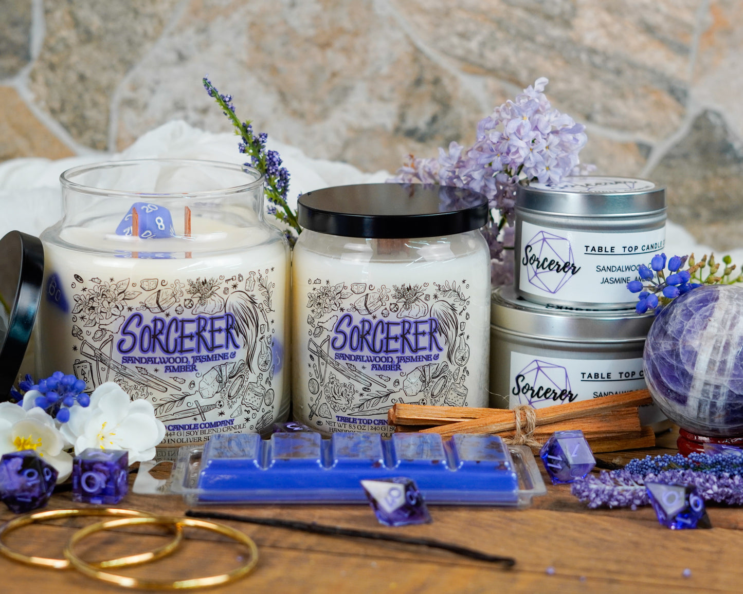 A full product lineup of the "Sorcerer" candle collection by Table Top Candle Company, including large and small jars, tins, and a snap bar. Surrounded by purple and white flowers, lavender sprigs, blue dice, and crystals for a magical, mystical vibe.
