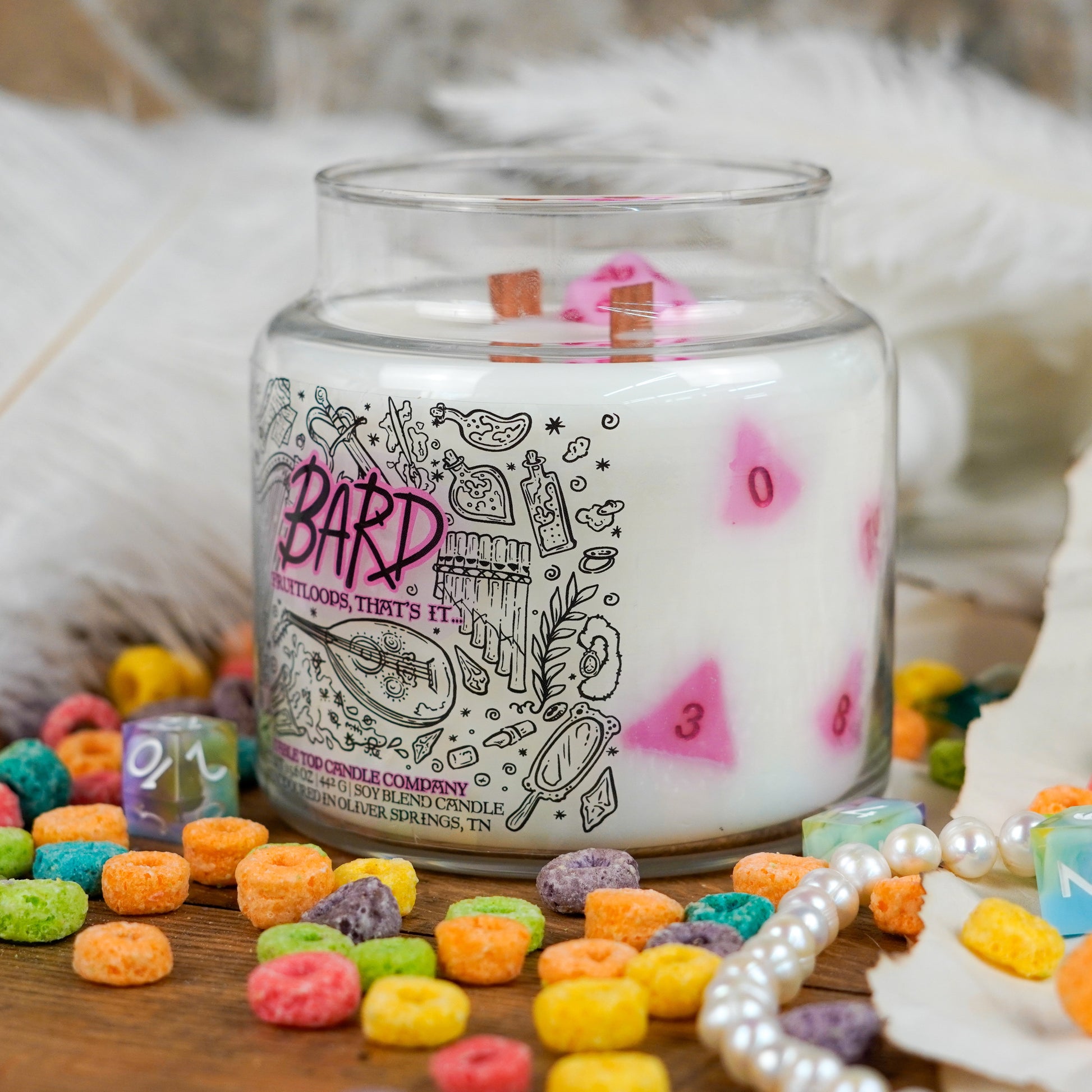 Side view of the "Bard" soy candle showcasing pink polyhedral dice embedded in the wax. The candle is set among cereal pieces, white feathers, and tabletop RPG dice for a whimsical, colorful display.