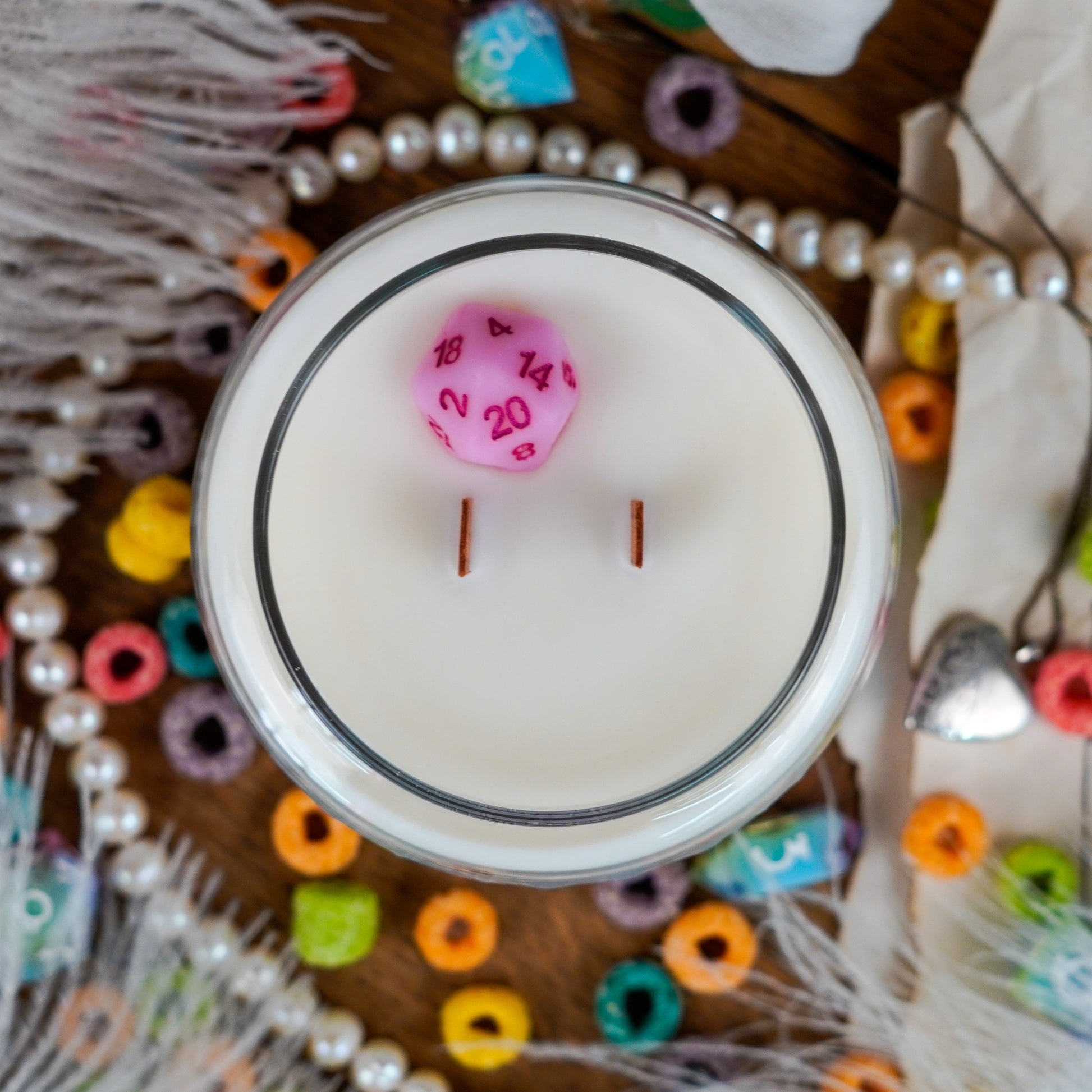 Top view of the "Bard" candle with two wooden wicks and a visible pink twenty-sided die (D20) embedded in the wax. The candle is surrounded by pearls, cereal, dice, and fantasy props.