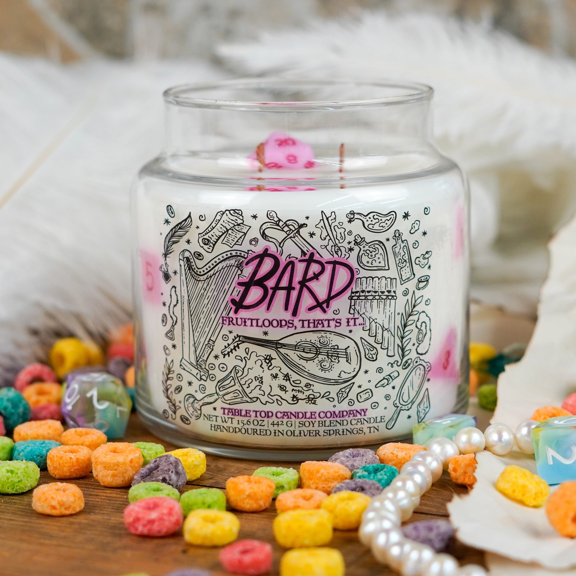 A fantasy-themed soy candle labeled "Bard – Fruitloops, That's It" with illustrations of instruments and musical notes. A pink polyhedral die is embedded in the wax. The candle is surrounded by feathers, pearl strands, and colorful cereal pieces.