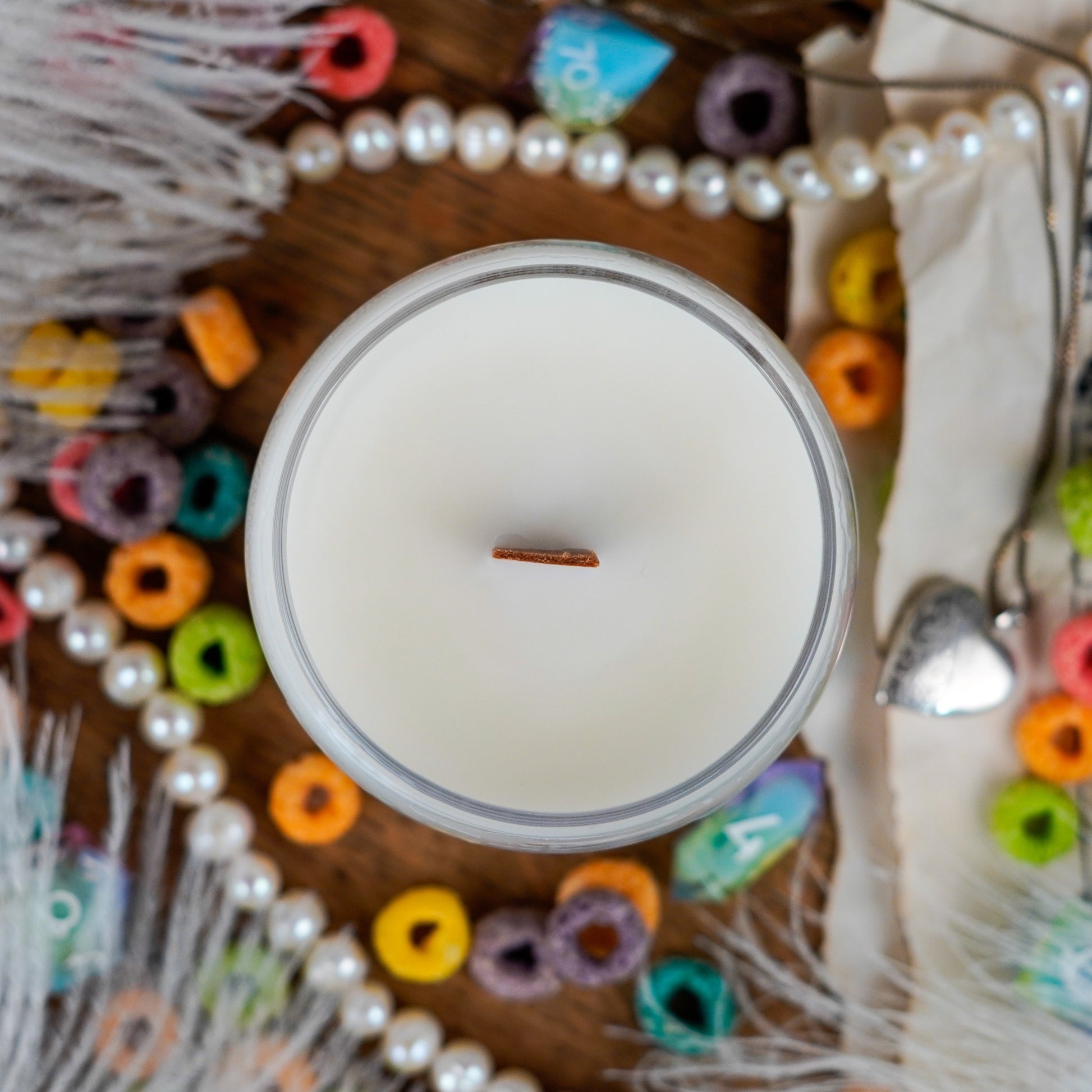 Top-down view of the "Bard" candle showing a single wooden wick centered in smooth, creamy wax. The surrounding scene includes colorful cereal, pearl strands, and feathers.
