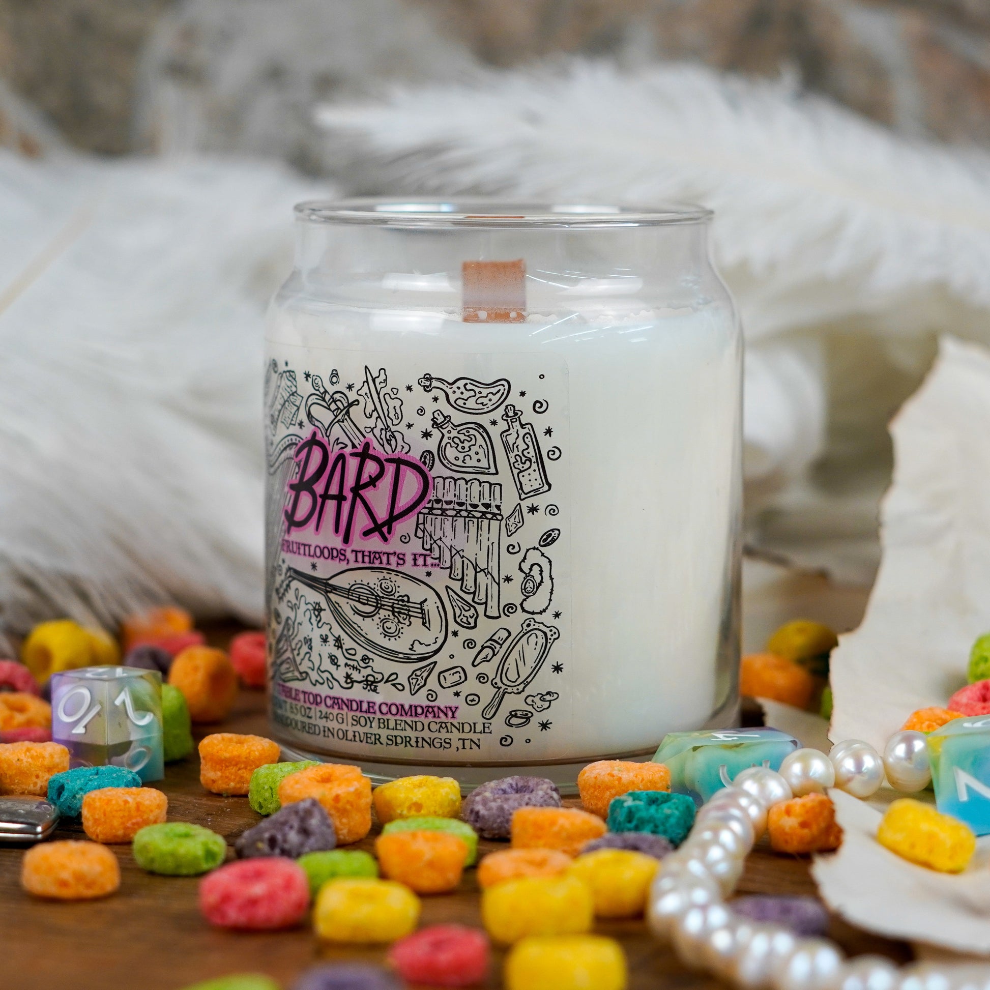 Angled view of the "Bard" candle highlighting its label and wooden wick. Set in a whimsical flat lay with fruit loop cereal, a pearl necklace, and feathers.