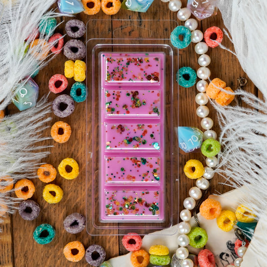A vibrant pink snap bar wax melt with glittery chunks, labeled as part of the "Bard" collection. The melt is displayed with playful accessories including cereal loops, feathers, pearls, and colorful dice.