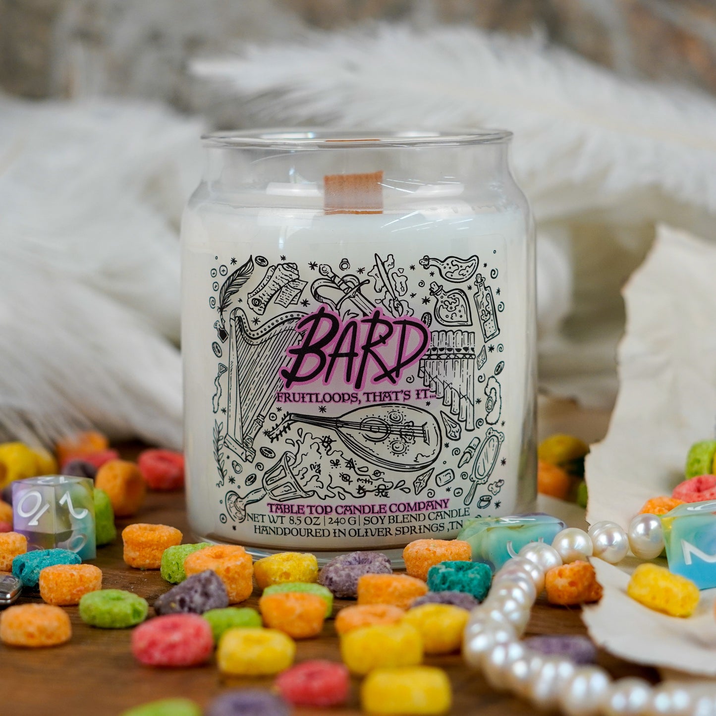 A smaller version of the "Bard" candle from Table Top Candle Company, centered in a bright, playful scene with rainbow cereal, dice, pearls, and white feathers. The candle has a wooden wick and musical illustrations on the label.