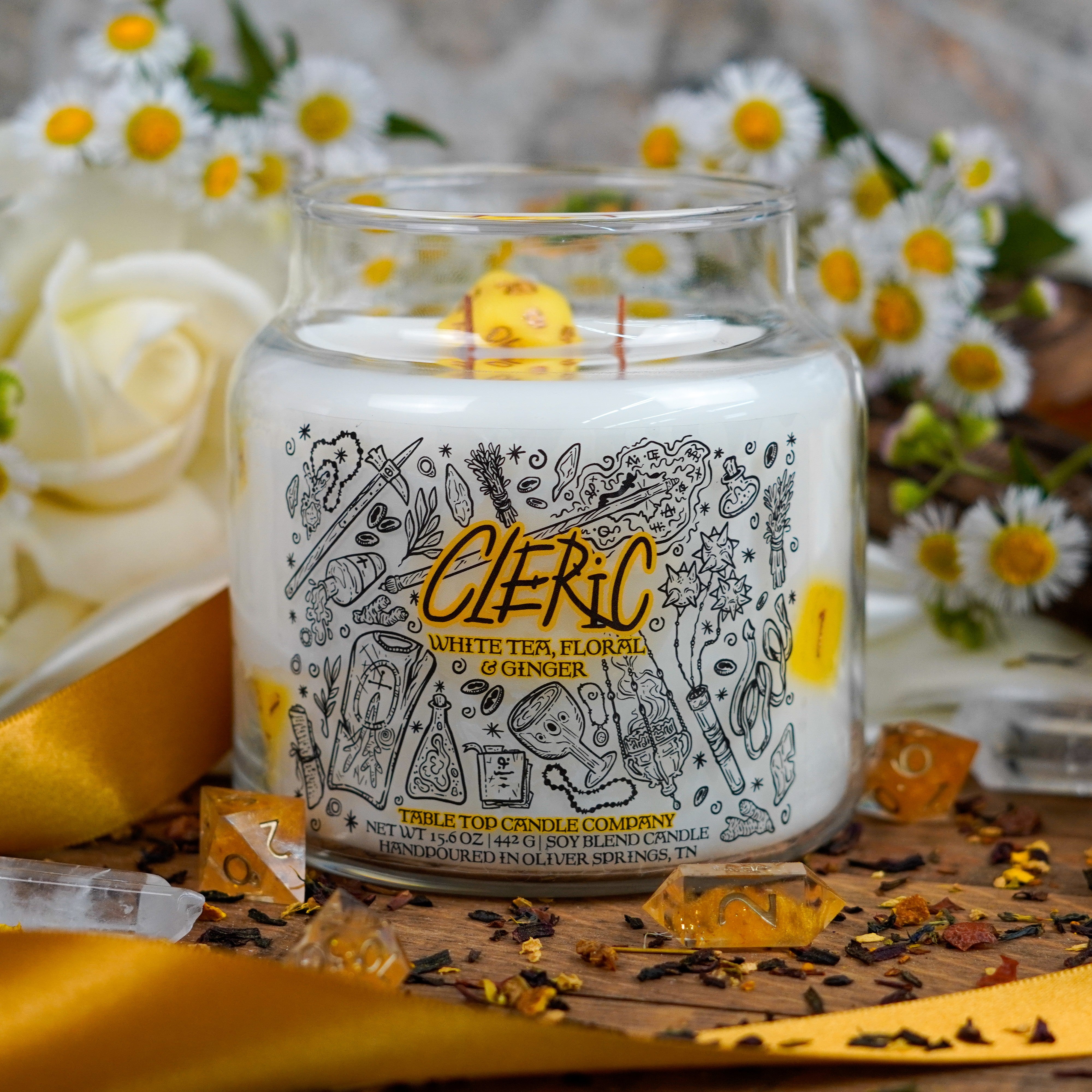 A fantasy-themed candle labeled "Cleric – White Tea, Floral & Ginger" from Table Top Candle Company. The white soy candle includes yellow polyhedral dice and is surrounded by white roses, chamomile flowers, loose tea, and satin ribbon.