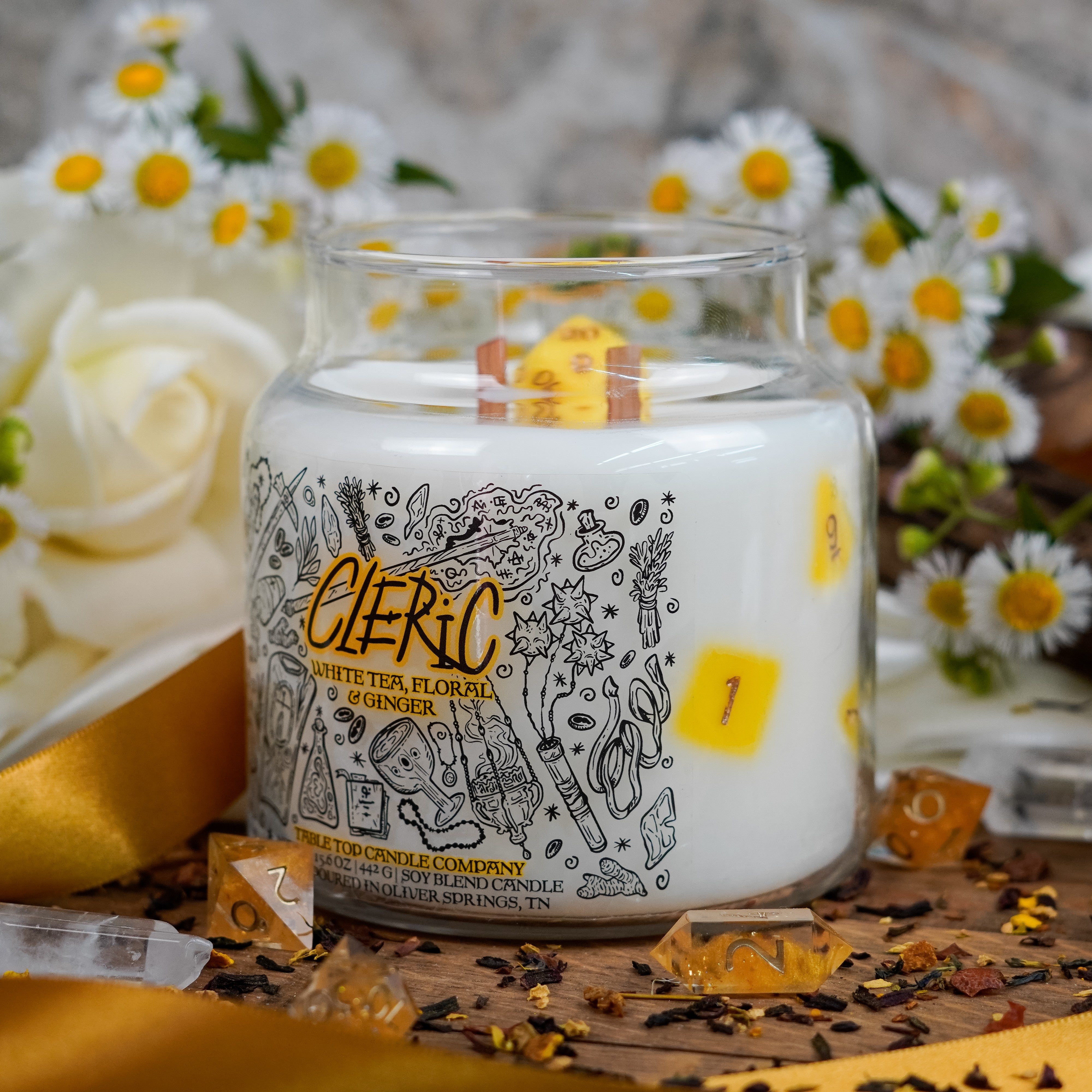 Side angle of the "Cleric" candle featuring embedded yellow dice. The label is decorated with magical and religious symbols. The candle is set among white and yellow flowers, gold ribbon, tea leaves, and quartz crystals.