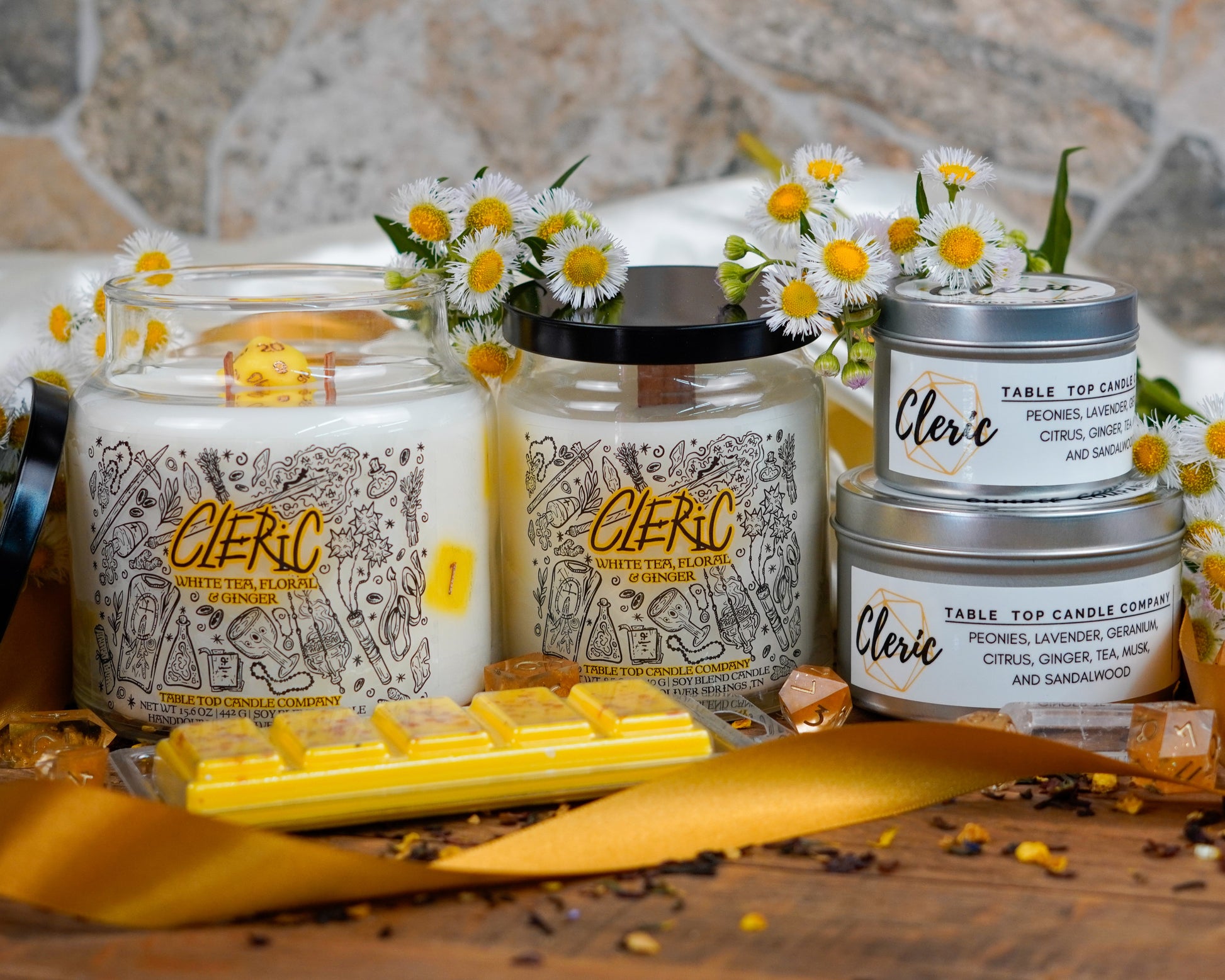 A full product lineup of the "Cleric" candle collection, featuring jar candles, tins, and snap bars. The scene is accented with gold ribbon, chamomile flowers, yellow dice, and tea leaves for an elegant and healing vibe.