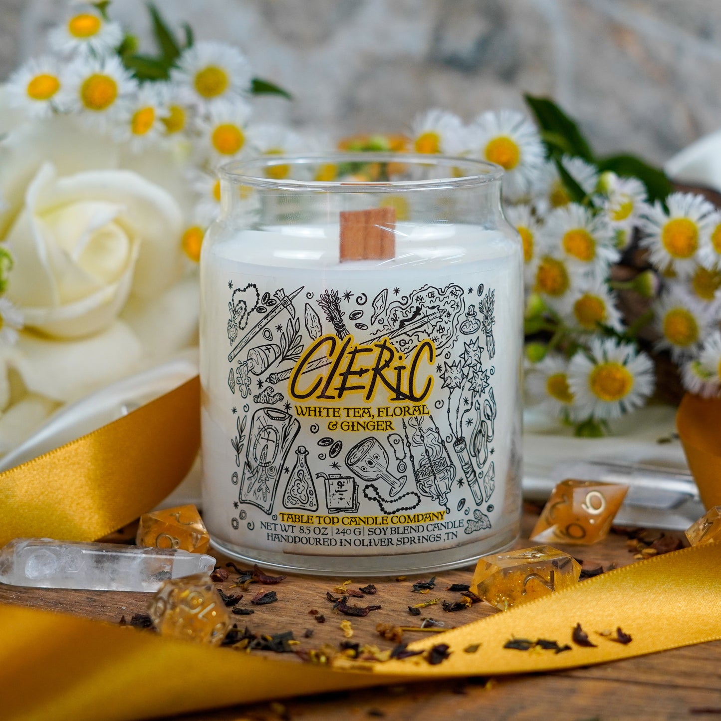 A smaller "Cleric" candle with a wooden wick, showcased in a peaceful floral arrangement of daisies and white roses. Surrounded by gold ribbon, crystals, and dried tea ingredients.