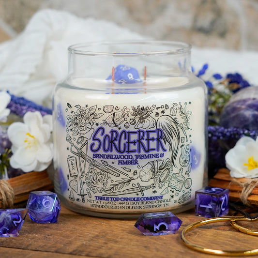 Front-facing view of the "Sorcerer" candle showcasing its magical-themed label with illustrations of potions, staffs, and spellbooks. A blue polyhedral die is embedded in the wax, and the scene is styled with amethyst tones and floral elements.