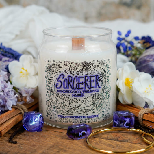 A smaller version of the "Sorcerer" candle, centered among lavender, white blossoms, and purple dice. The soy candle features a wooden wick and fantasy illustrations in shades of violet and black.